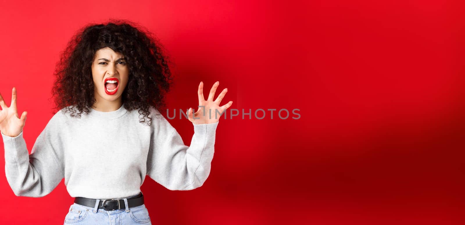Scary woman trying to scare you, shouting and showing animal claws gesture, screaming at person, standing against red background.