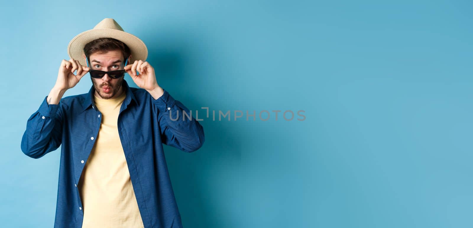 Portrait of tourist takes-off sunglasses and saying wow, staring impressed at camera, checking out cool thing, standing in summer hat on blue background.