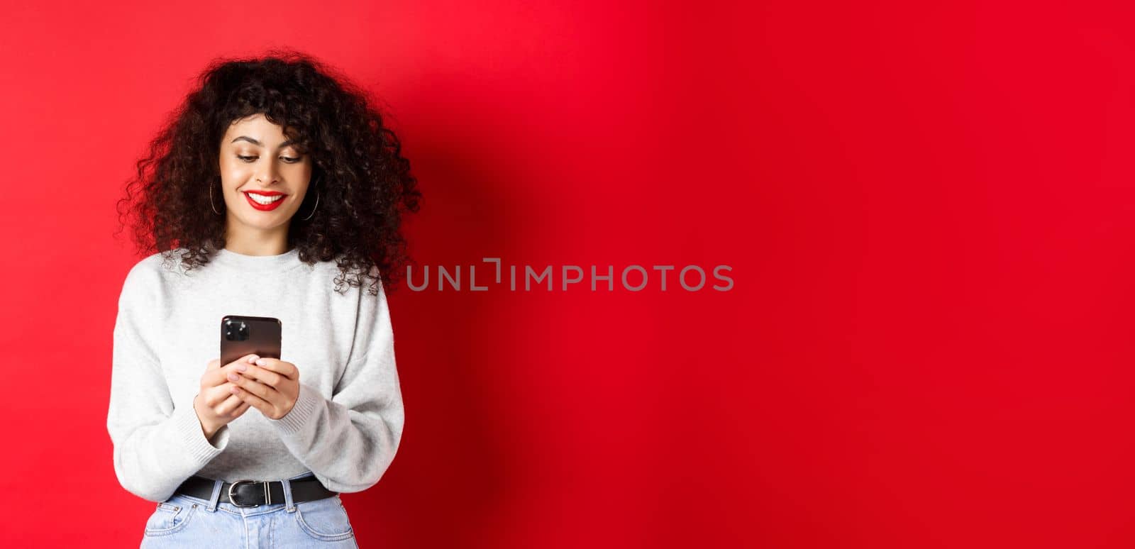 Smiling european woman reading mobile phone screen, texting a message on smartphone, standing in sweatshirt against red background.