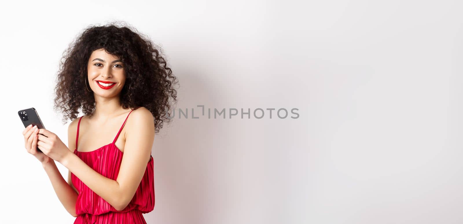 Happy elegant woman in red dress writing message, using smartphone and smiling at camera, standing against white background by Benzoix