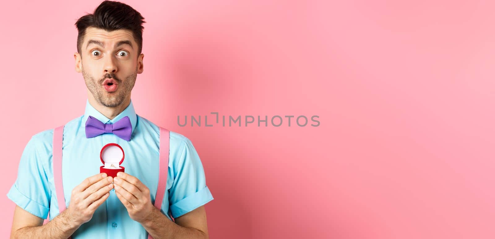 Valentines day. Funny man with moustache and bow-tie, looking excited and showing engagement ring, asking girlfriend to marry him, standing over pink background by Benzoix