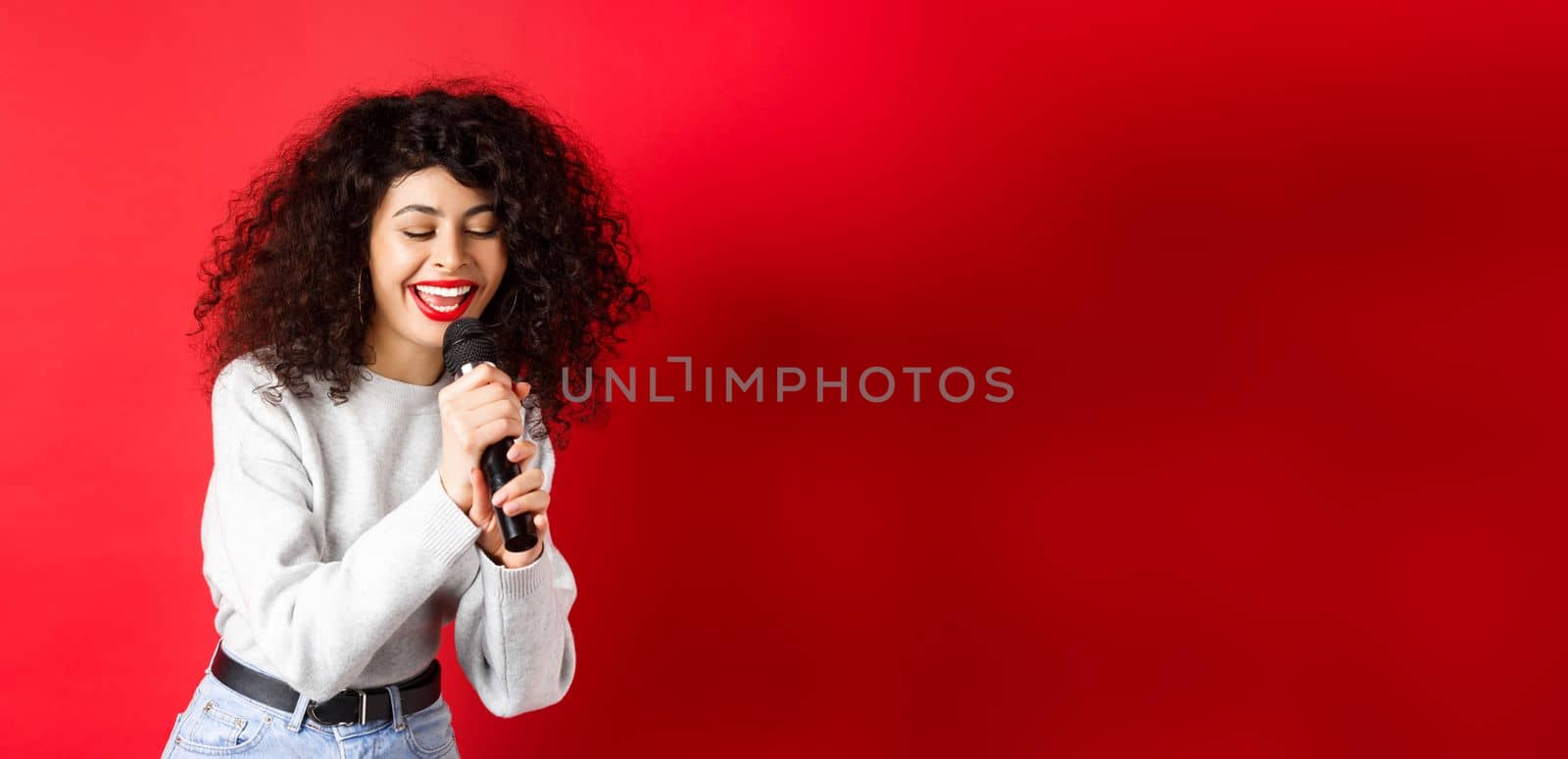 Hobbies and leisure concept. Beautiful woman singing in microphone and smiling, having fun at karaoke, standing on red background by Benzoix