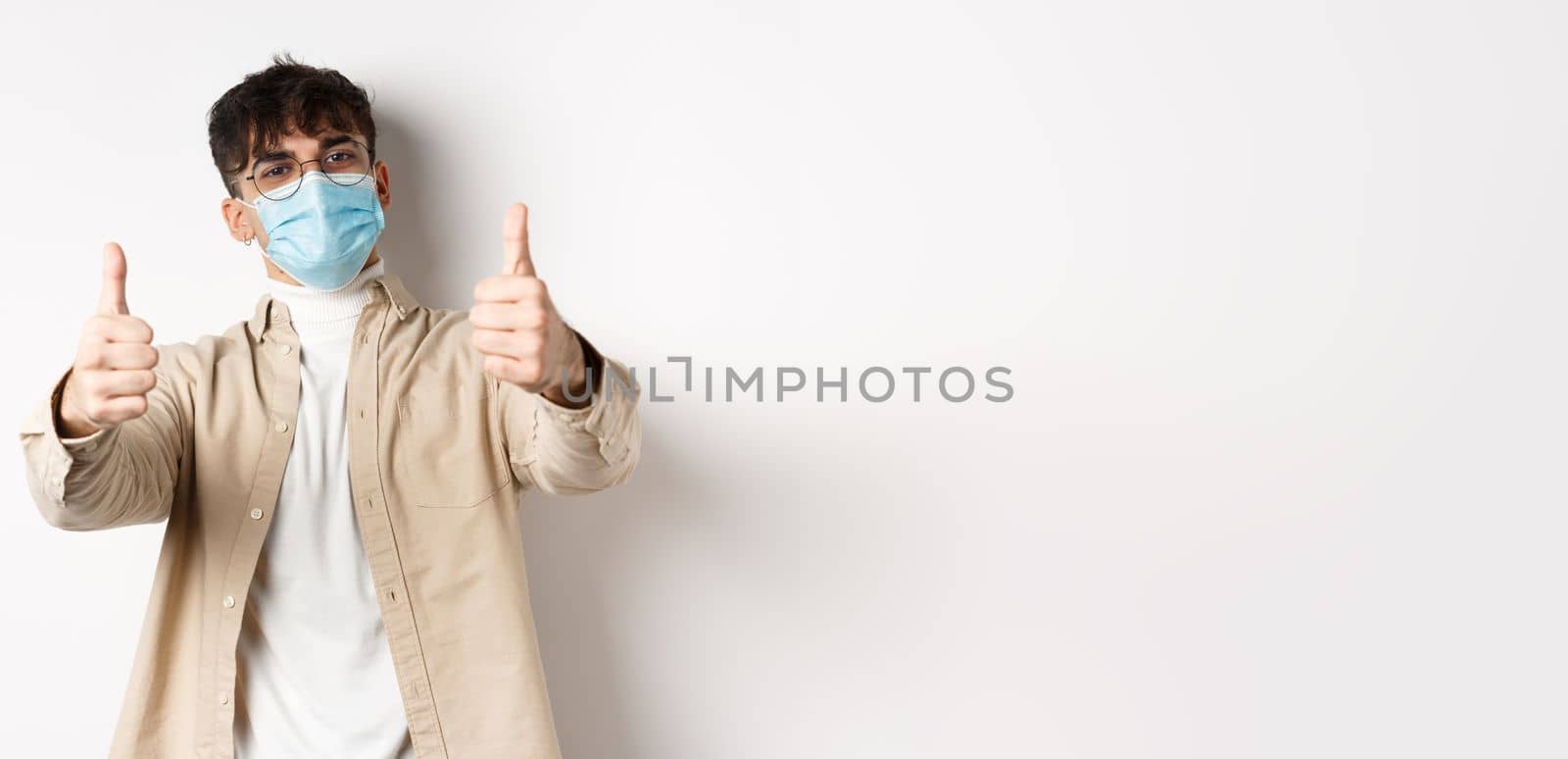 Coronavirus, health and real people concept. Smiling guy in medical mask showing thumbs up, wearing glasses, standing on white background.