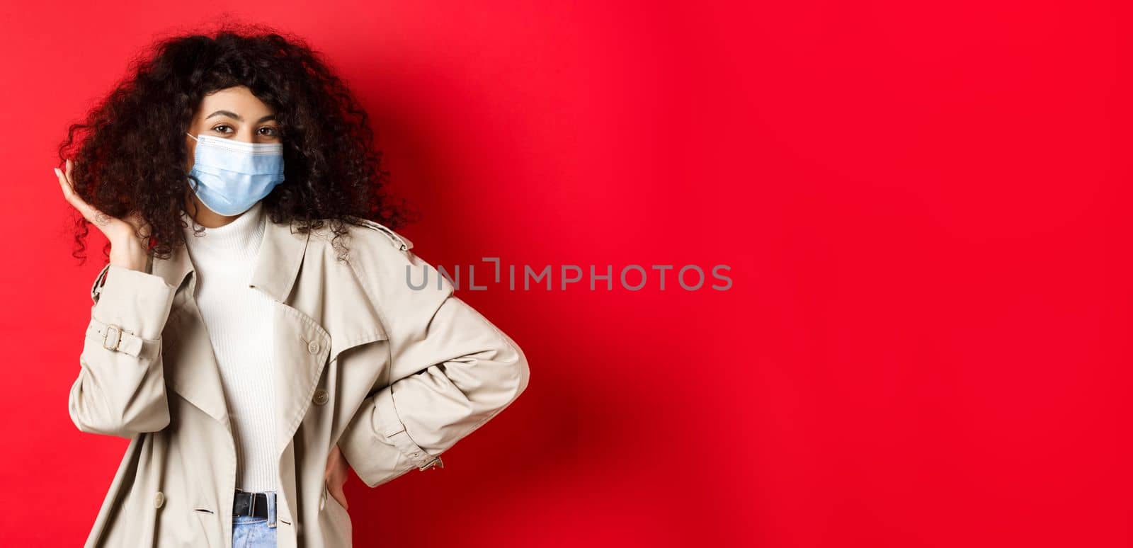 Covid-19, pandemic and quarantine concept. Stylish coquettish woman in medical mask and trench coat, fixing her curly hairstyle and smiling, red background.