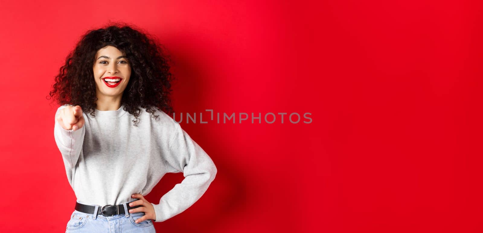Smiling cheerful woman with curly hairstyle, pointing fingers at camera, inviting you, choosing someone, standing on red background.
