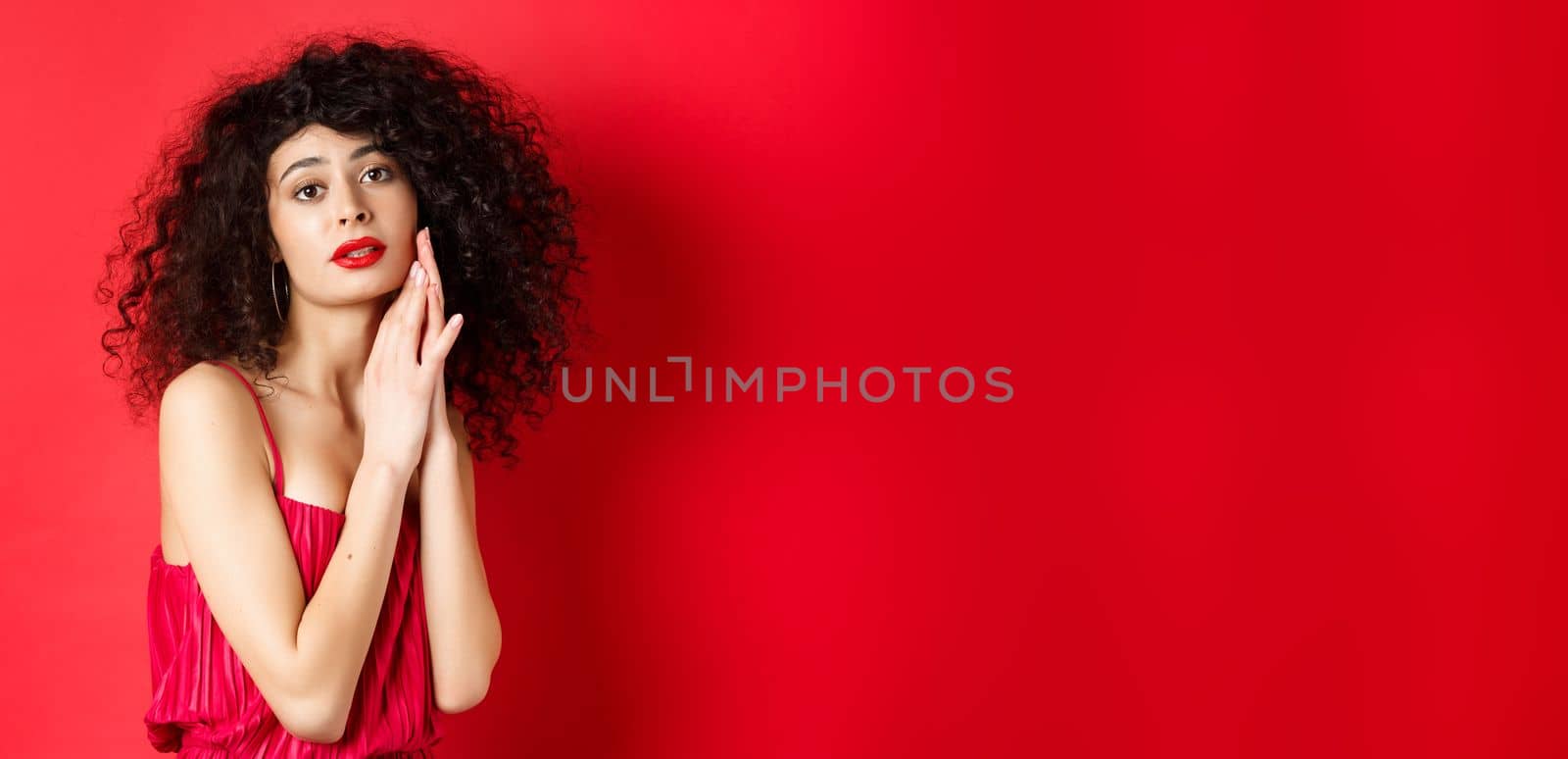 Romantic sensual woman with curly hair, wearing evening dress, posing seductive on red background.
