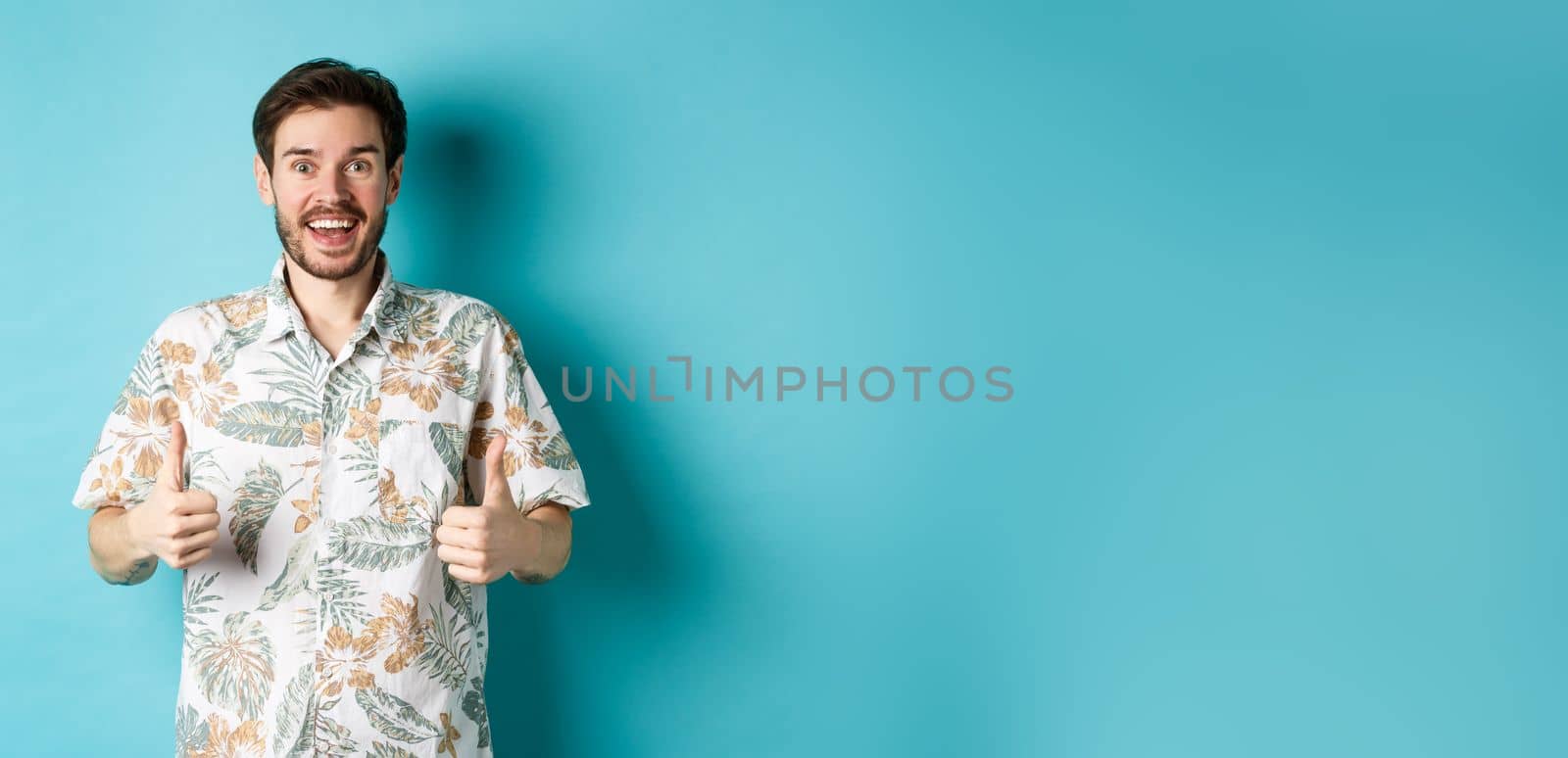 Cheerful guy showing thumbs up on vacation, praising travel agency, standing in summer shirt on blue background.