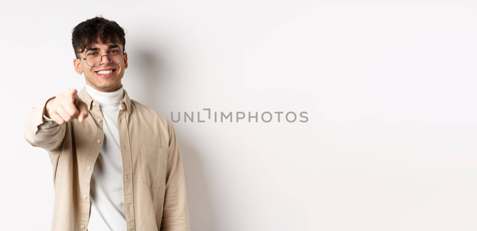 Handsome modern guy in glasses pointing at camera, smiling and choosing you, recruiting or inviting to event, standing on white background.