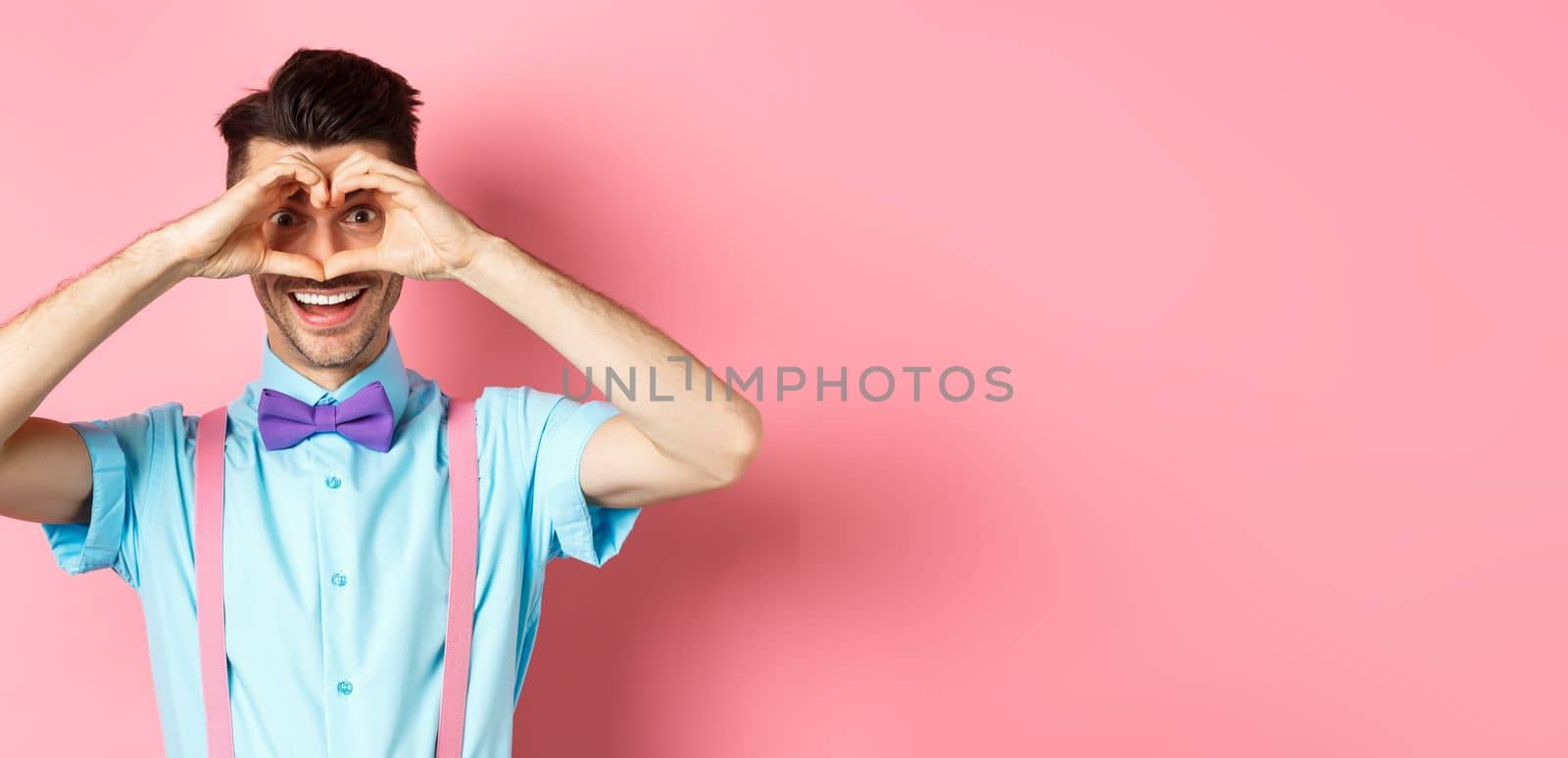 Happy Valentines day. Young man making hand heart and looking through it at camera, feeling romantic with lover, standing over pink background.