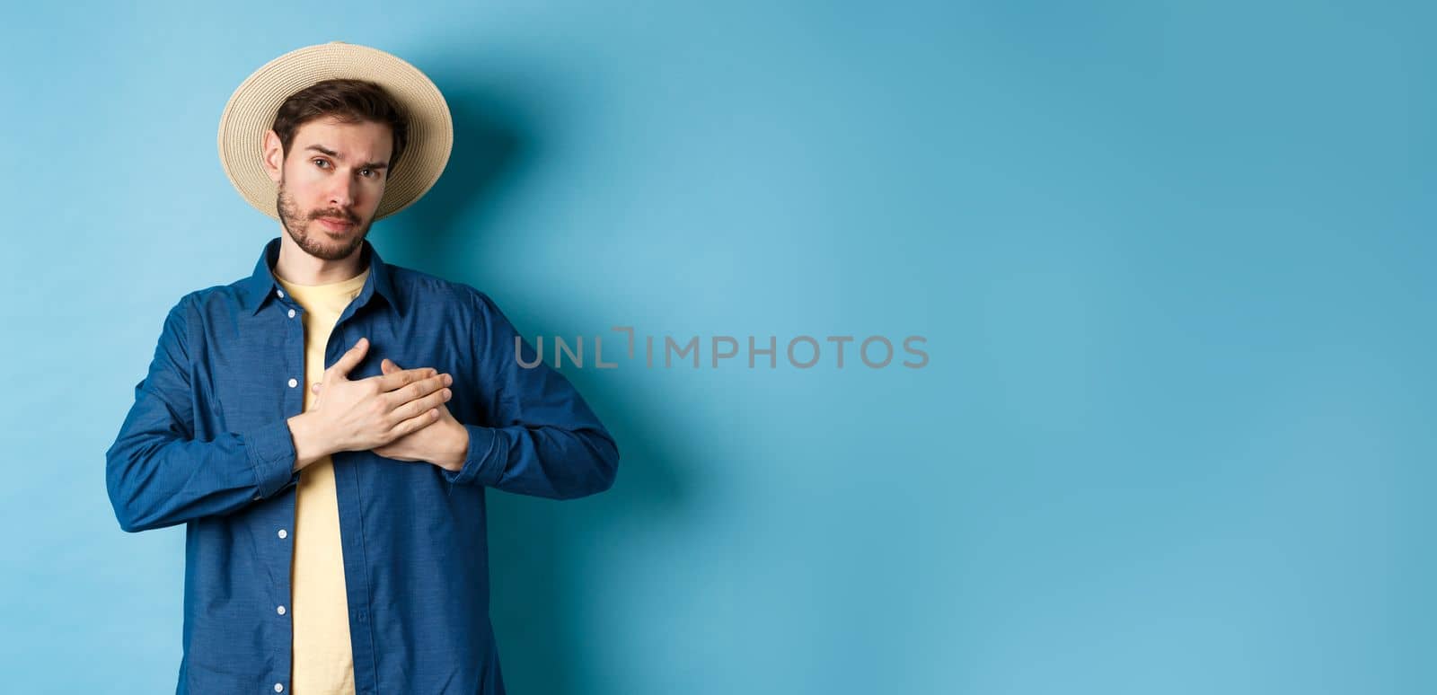 Heartfelt young woman in tourist hat, holding hands on heart and looking thankful and serious, standing on blue background by Benzoix