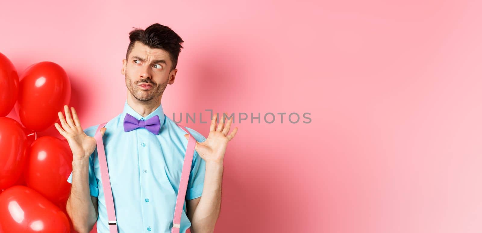 Valentines day concept. Pensive young man in bow-tie, raising hands up and looking left while thinking, making decision, standing on romantic pink background and heart balloons.
