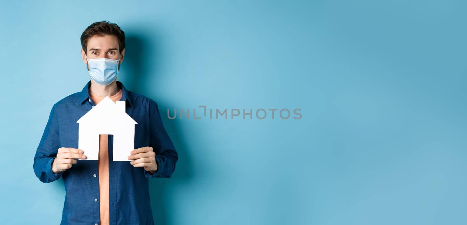 Real estate and quarantine concept. Happy young man in medical mask showing house cutout, buying property, standing on blue background by Benzoix