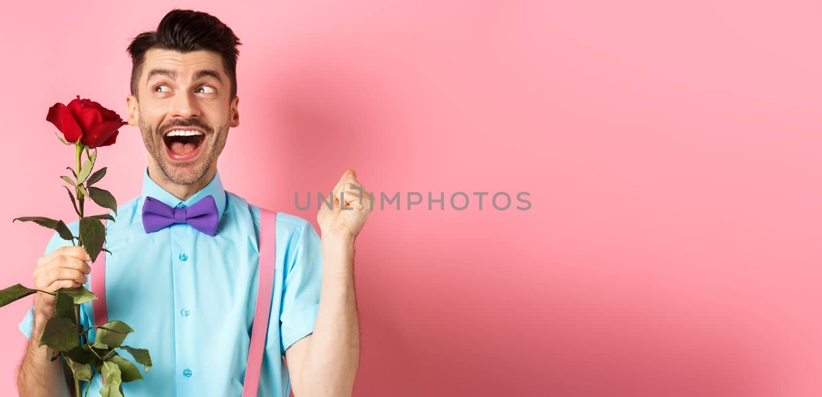 Romance and Valentines day concept. Cheerful man in bow-tie screaming from happiness, holding red rose and jumping on date, standing over pink background.