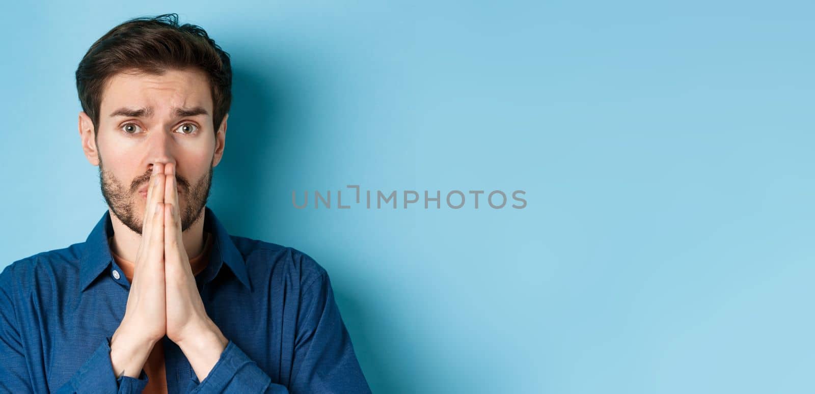 Close up of sad young man pleading, begging for help and say please, standing on blue background.