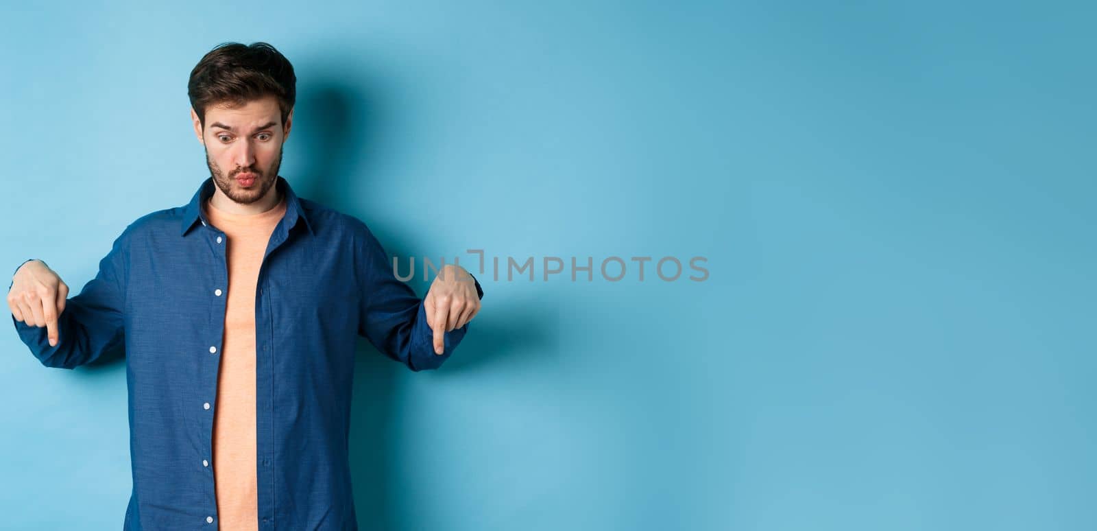 Image of shocked young man in casual clothes, looking and pointing down at something strange, standing on blue background by Benzoix