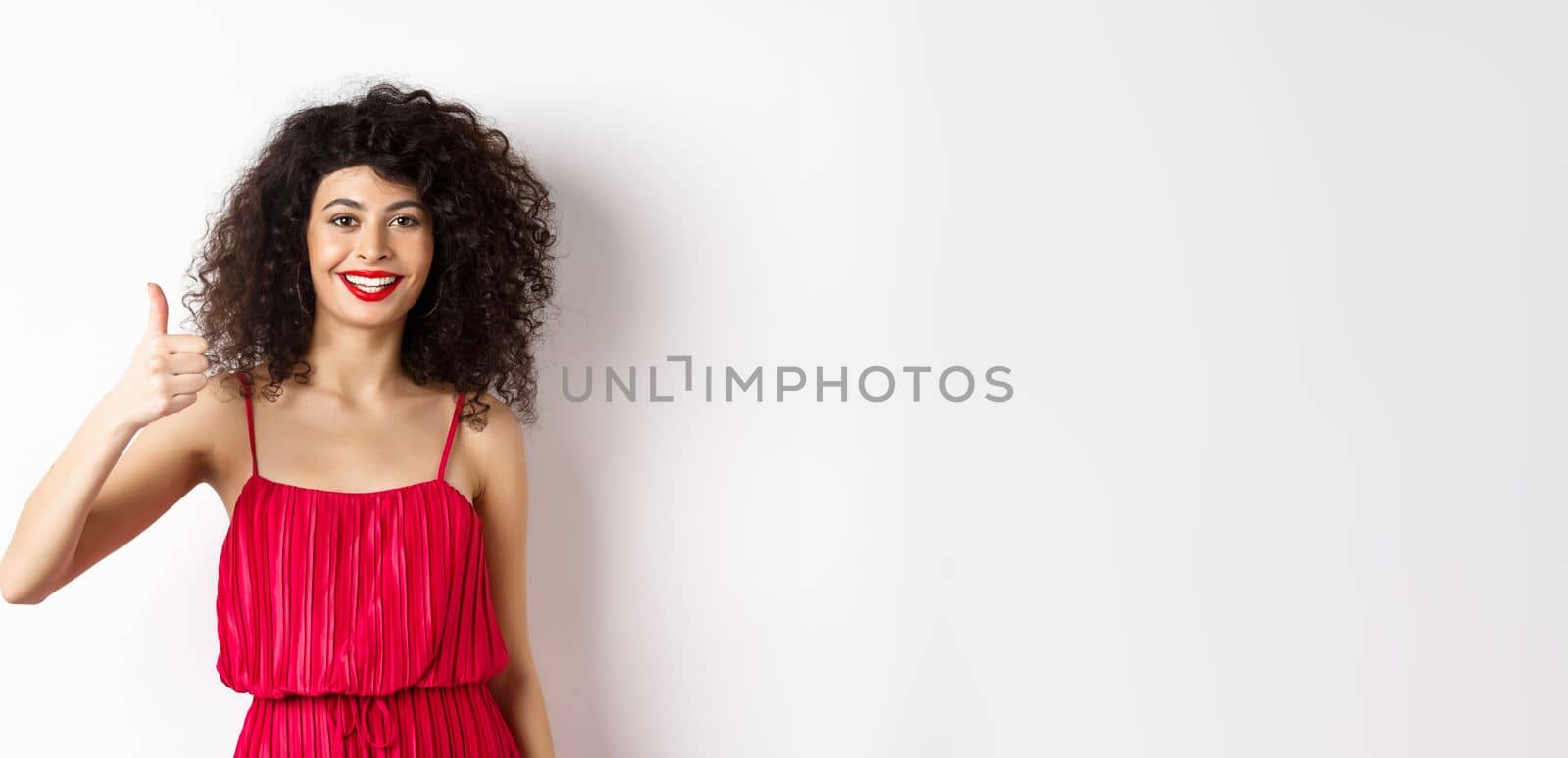 Cheerful woman with red lips and elegant dress, showing thumb up and smiling, recommending product, standing over white background.