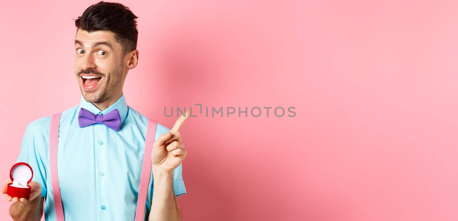 Valentines day. Funny guy with moustache and bow-tie, showing engagement ring and pointing finger at upper right corner, standing on pink background.