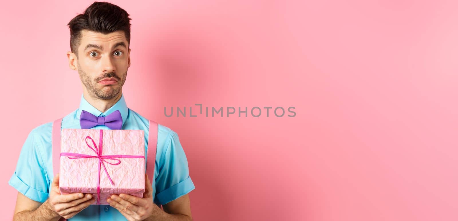 Holidays and celebration concept. Attractive young man with moustache, wearing festive outfit with bow-tie, showing cute gift box and looking at camera, standing over pink background.