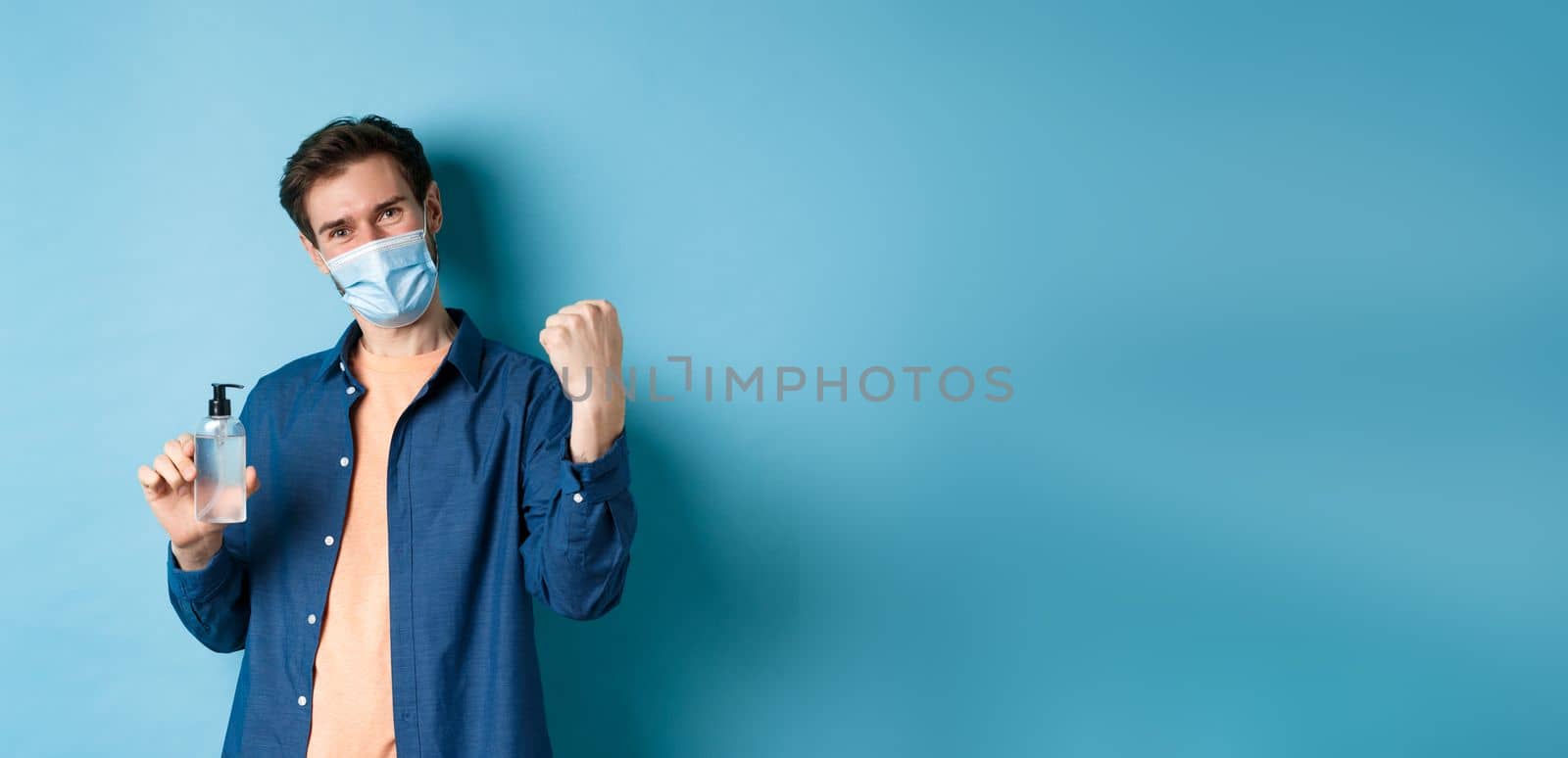 Coronavirus, quarantine and social distancing concept. Cheerful guy say yes and raise fist up while showing hand sanitizer, recommending product, blue background by Benzoix