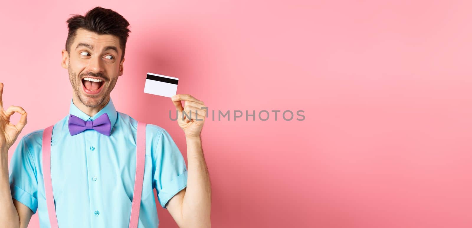 Cheerful man recommending great promo, showing OK sign and looking excited at plastic credit card, standing on pink background by Benzoix