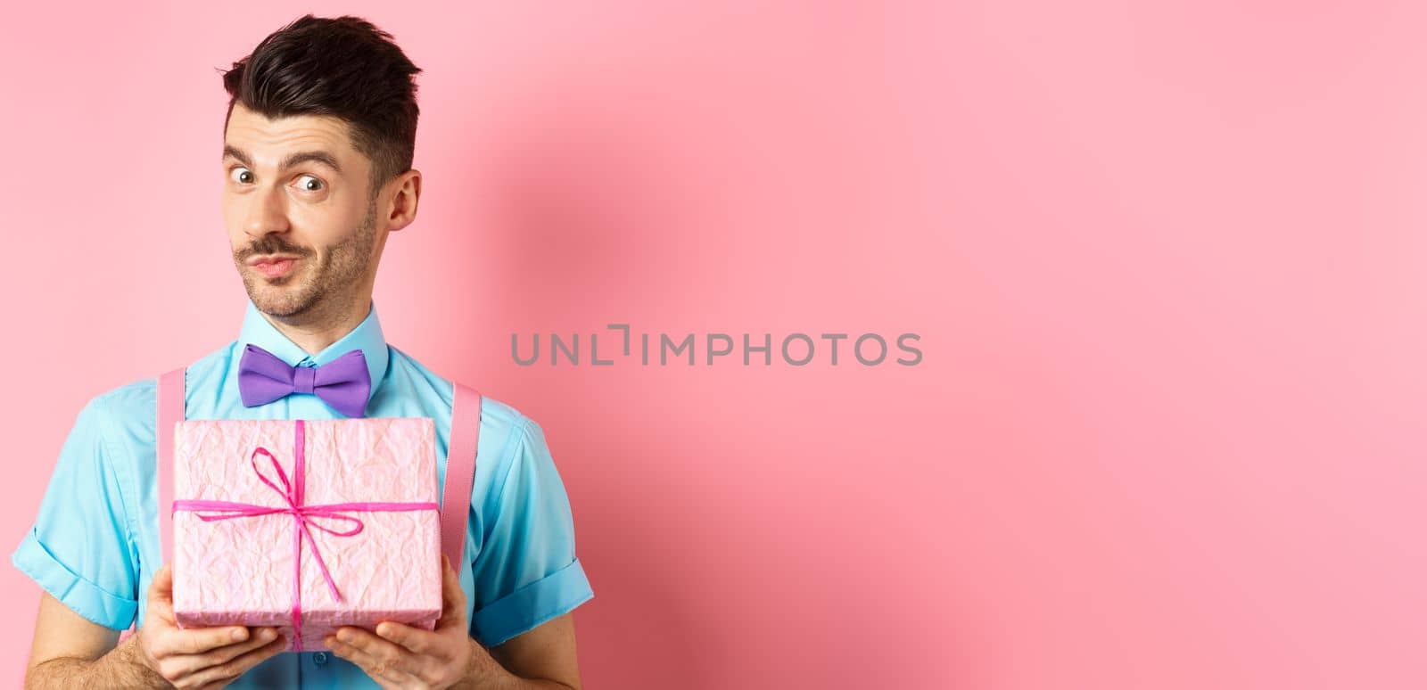 Holidays and celebration concept. Funny guy with moustache showing cute gift box wrapped in festive paper, looking at camera, presenting you, standing on pink background by Benzoix