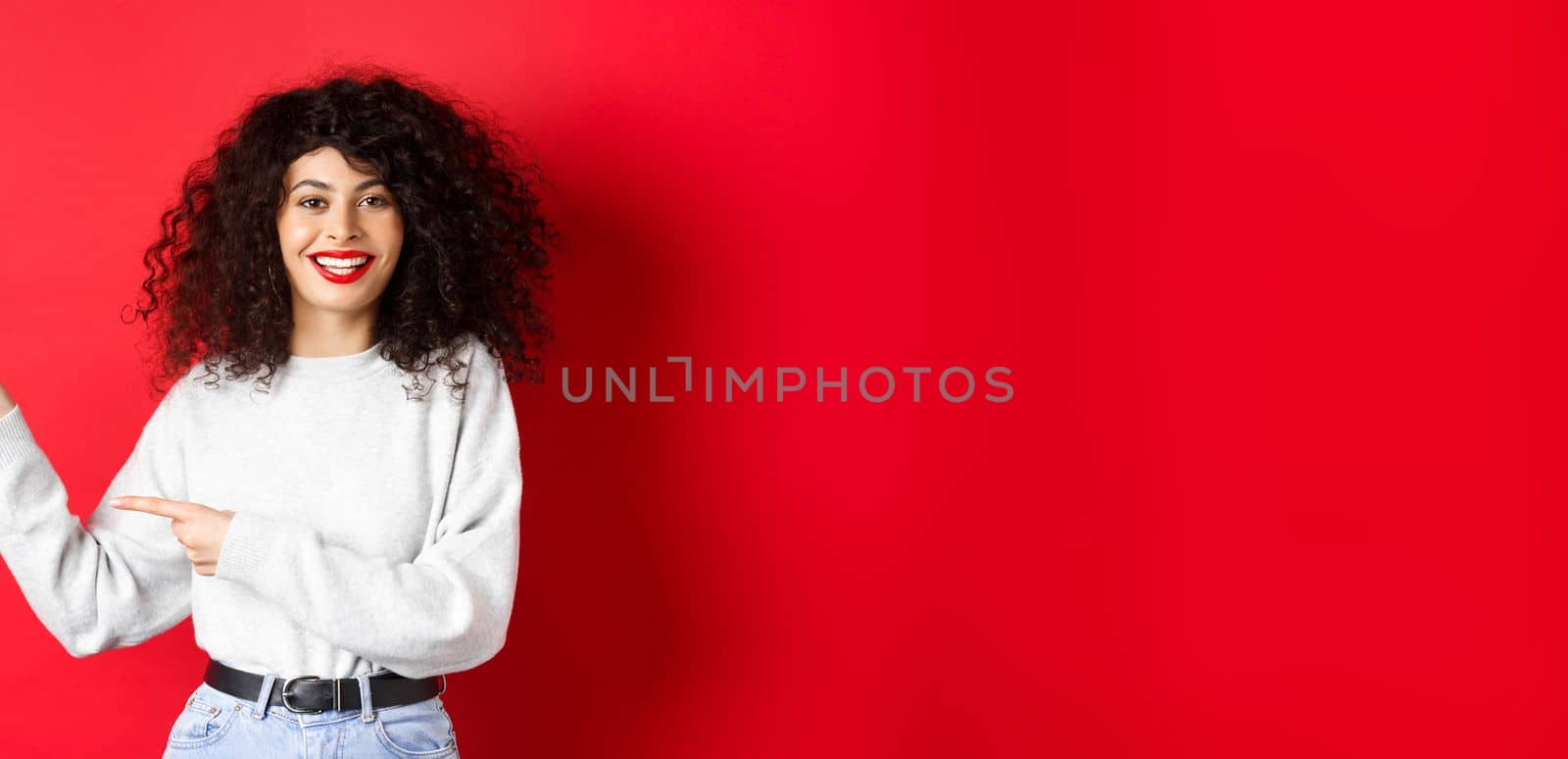 Smiling caucasian woman with curly hair and red lips, pointing fingers left at logo, showing promo offer, standing against studio background by Benzoix