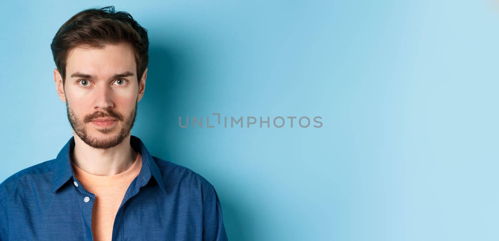 Close-up of young man with beard looking serious at camera, standing in casual shirt on blue background.