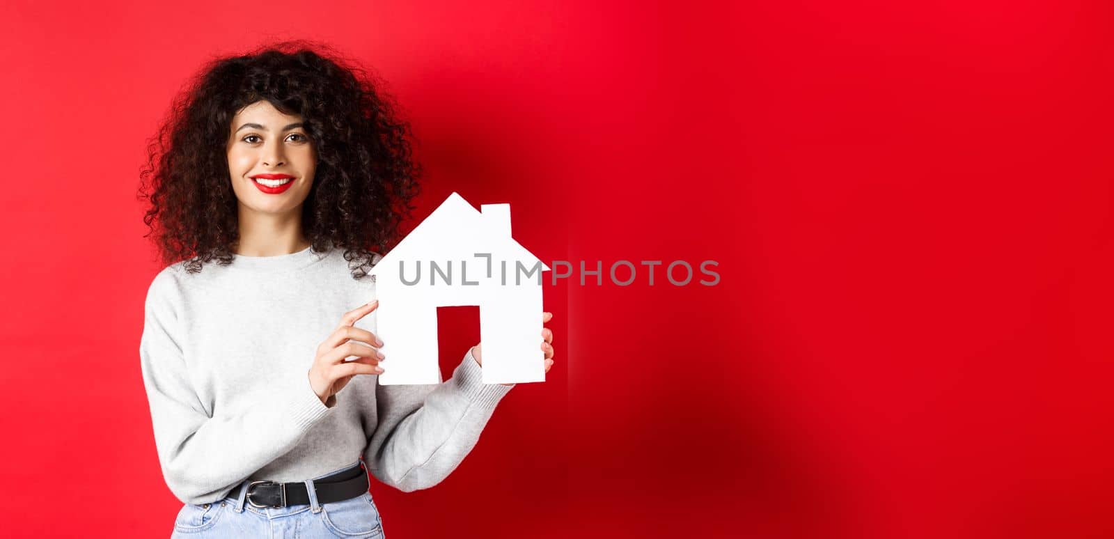 Real estate. Smiling caucasian woman with curly hair and red lips, showing paper house model, searching property, standing on red background by Benzoix