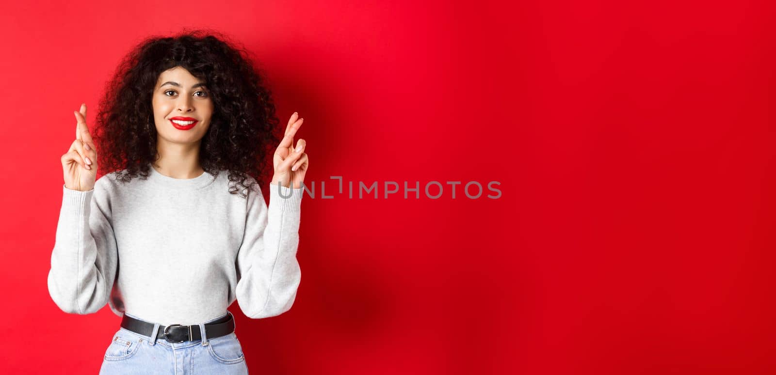 Hopeful young woman with red lips and curly hair, cross fingers for good luck and making wish, praying for dream come true, smiling excited, red background.