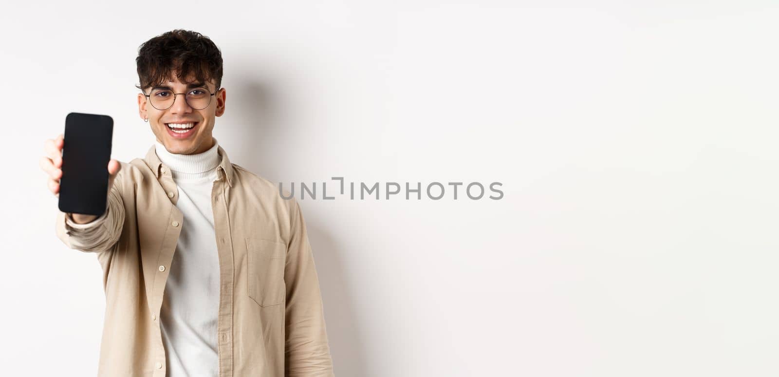 Handsome young man showing empty smartphone screen, standing on white background. Copy space