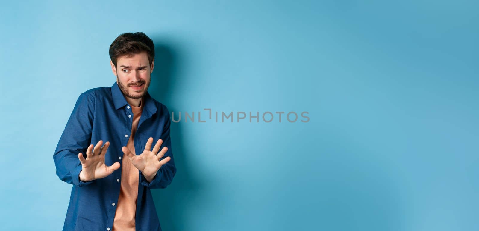 Man cringe and stay away from something nasty, looking aside and stretch out hands to reject bad deal, standing on blue background by Benzoix