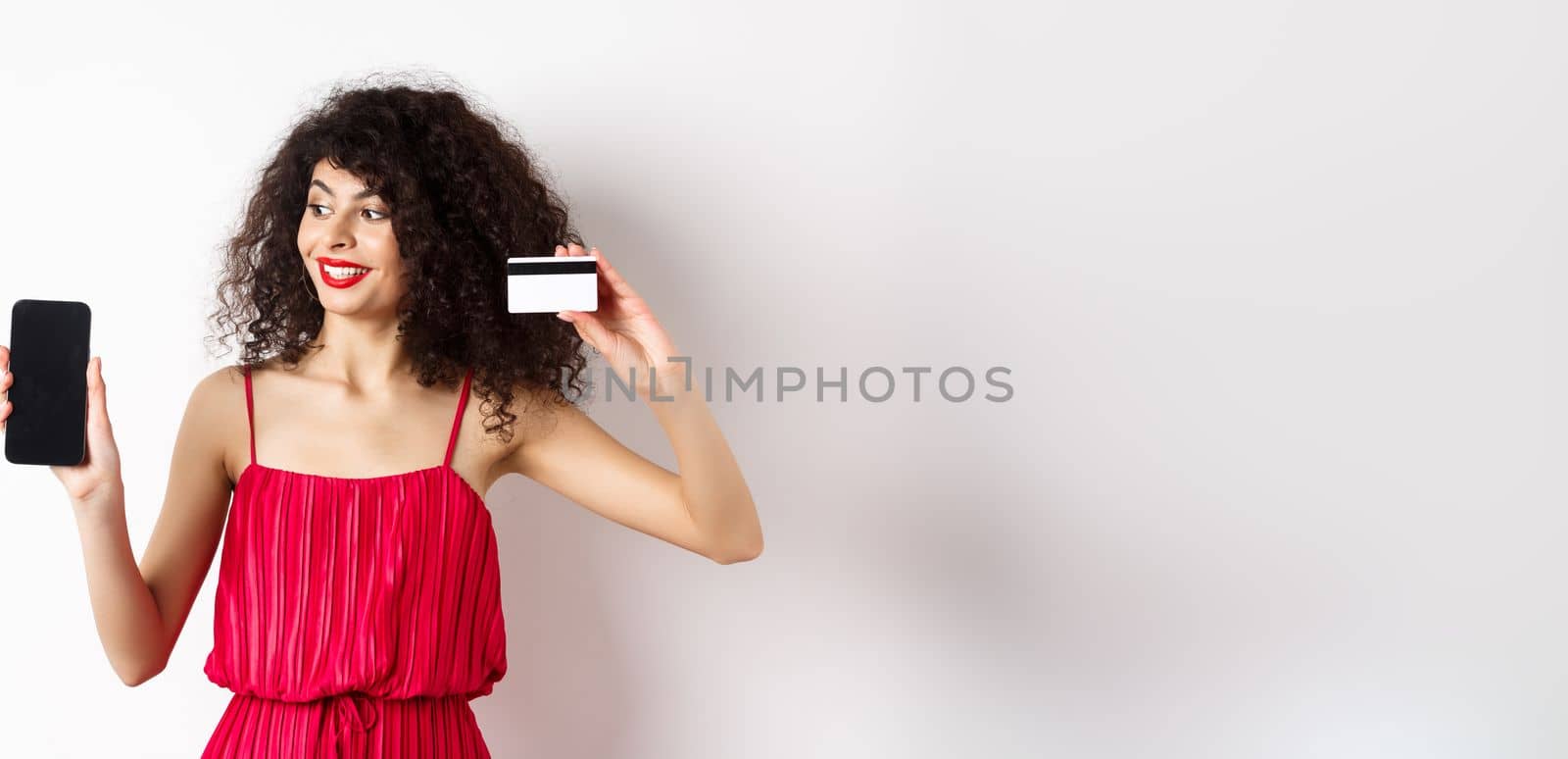 Online shopping concept. Elegant curly-haired woman in red dress showing plastic credit card and empty mobile phone screen, standing over white background.