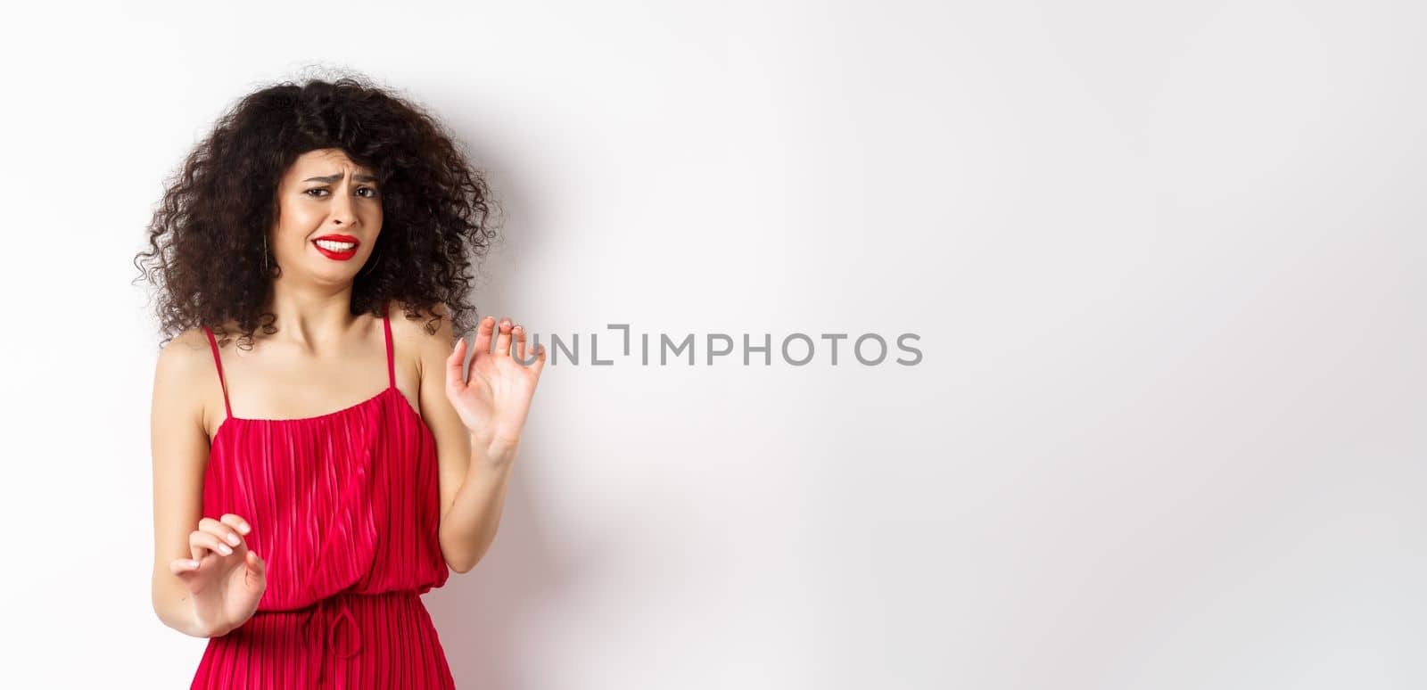 Young woman cringe of something creepy, grimacing and recoil from disgusting awful thing, standing in red dress on white background.