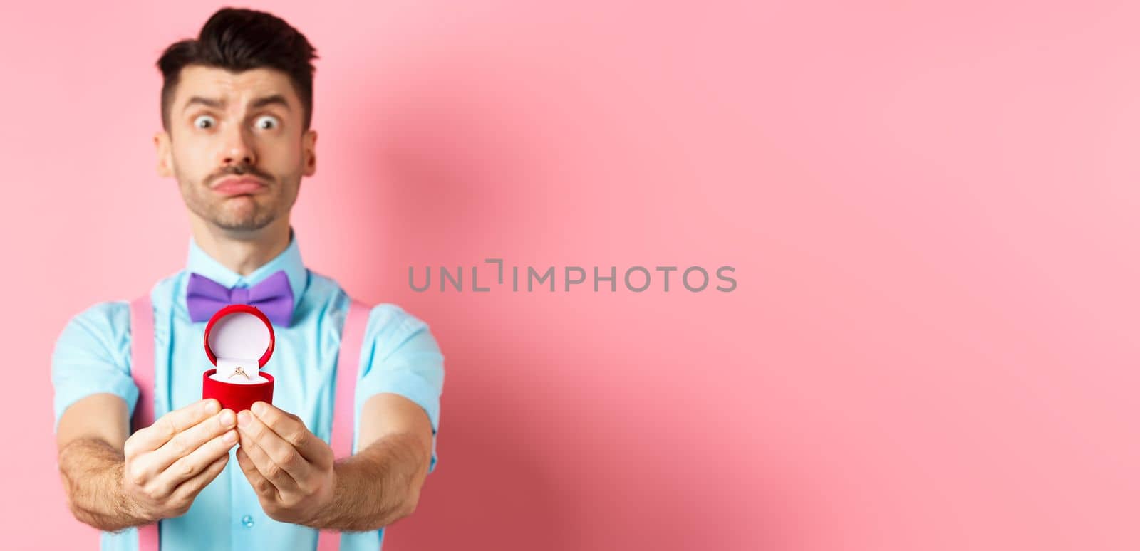 Valentines day. Nervous boyfriend waiting for girlfriend reply on marriage proposal, showing engagement ring and looking worried, standing over pink background by Benzoix