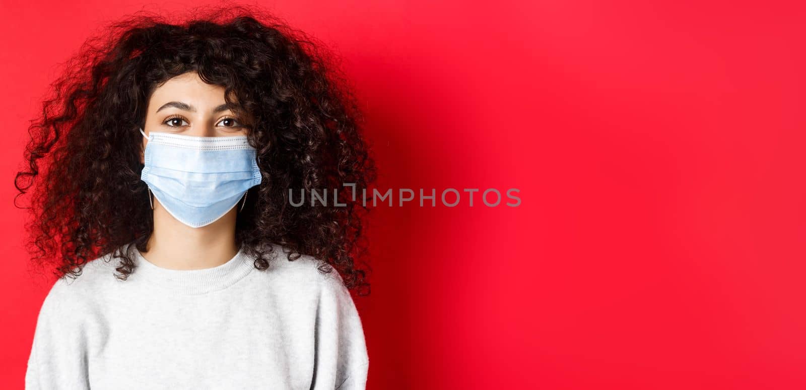 Covid-19 and pandemic concept. Close-up of modern young woman with curly hair, wearing medical mask from coronavirus, smiling at camera, red background.