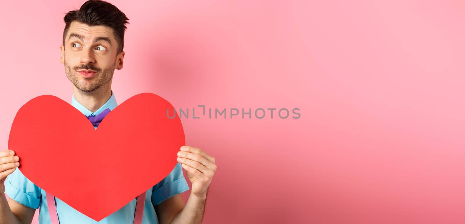 Valentines day concept. Dreamy romantic man looking left and showing big red heart cutout, standing on pink background.