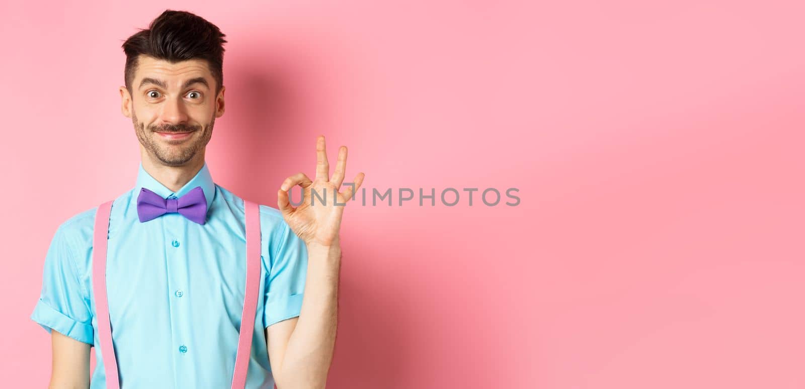 Smiling caucasian man in bow-tie and suspenders showing all okay gesture, recommending something cool, praising promo on pink background by Benzoix