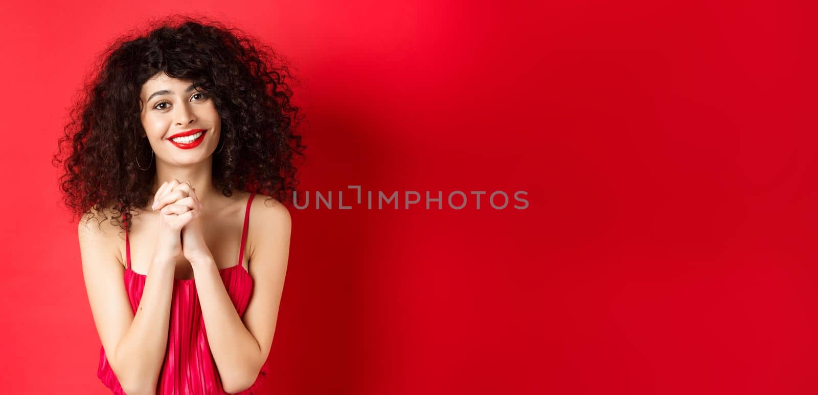 Image of beautiful curly-haired woman in red dress and makeup, saying thank you, looking grateful and smiling happy at camera, studio background.