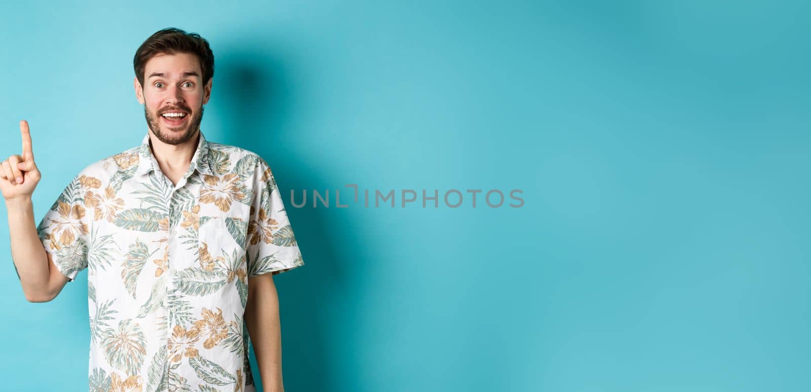 Excited guy in hawaiian shirt talking about vacation, looking amazed and pointing up, standing on blue background by Benzoix