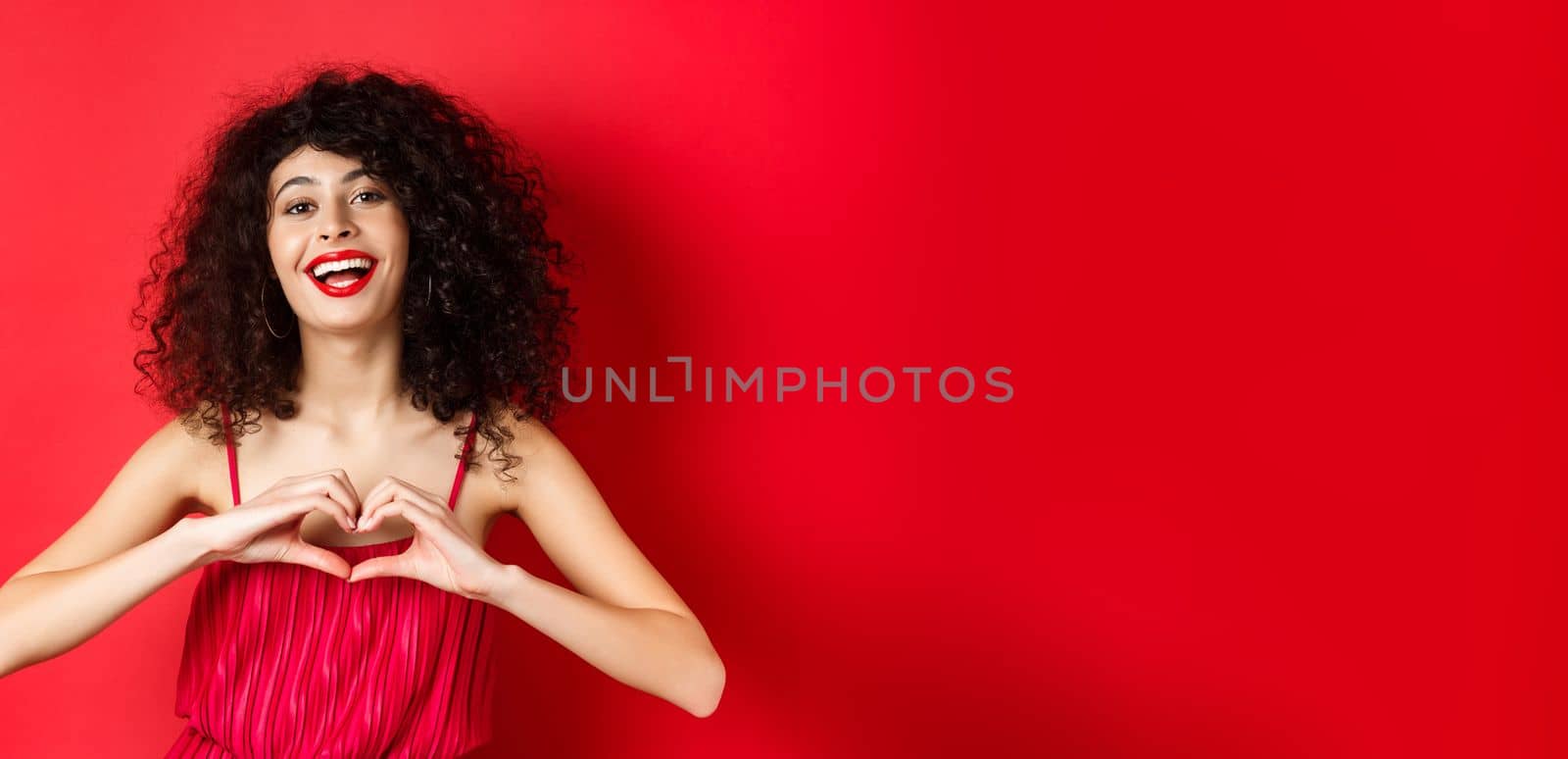 Beautiful woman with curly hair, red dress, showing heart symbol and smiling happy, studio background.