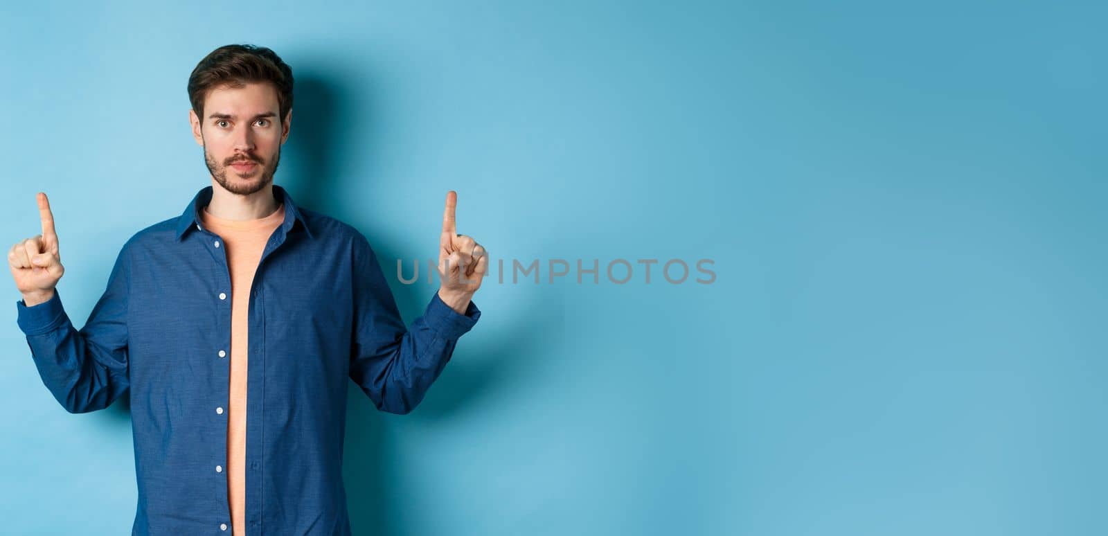 Serious caucasian man with beard pointing fingers up at empty space, showing logo and looking at camera, standing on blue background.