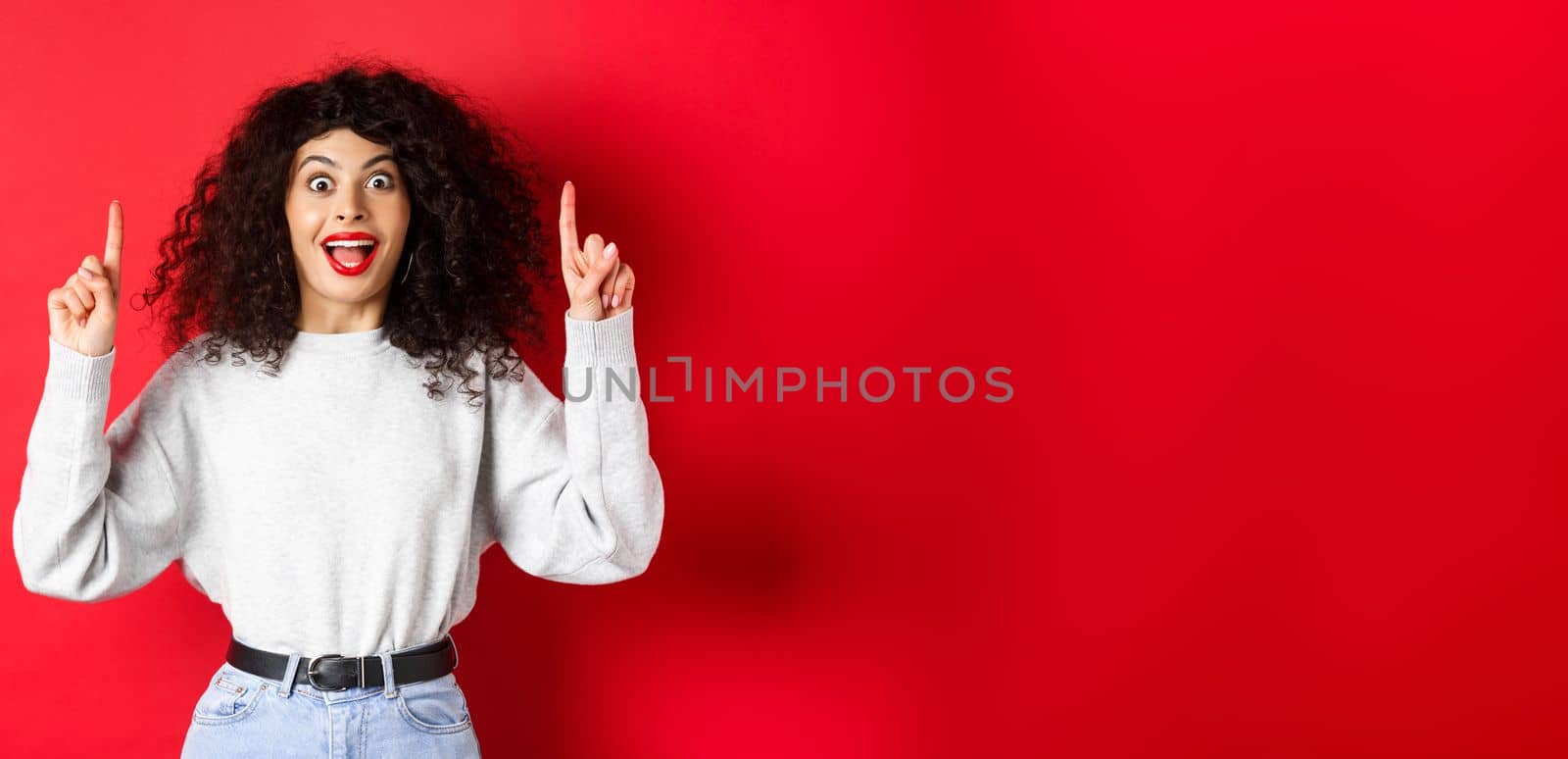 Excited modern girl showing promo, pointing fingers up and smiling amazed, telling big news, standing on red background.