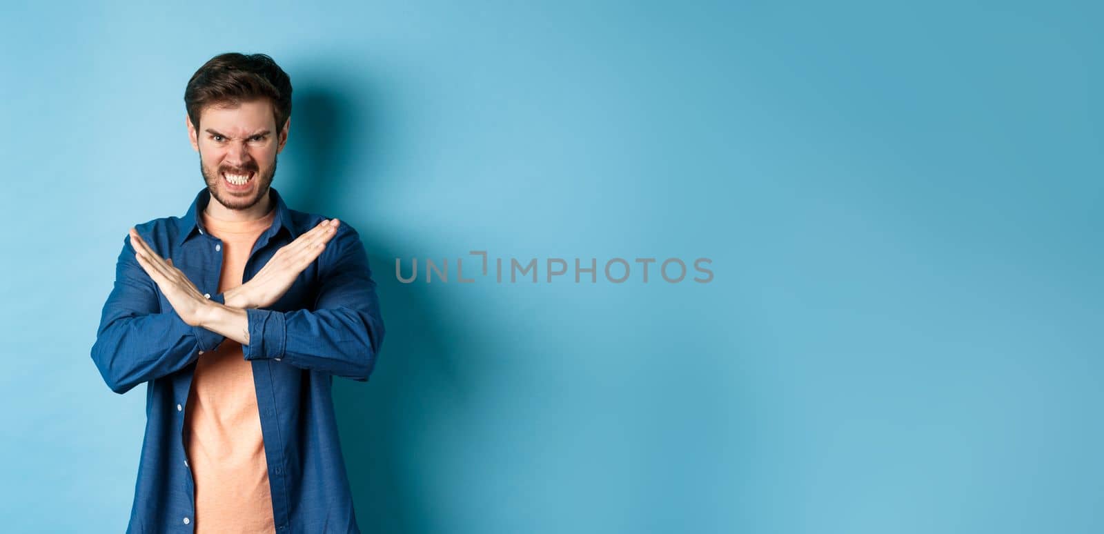 Angry young man frowning and clenching teeth outraged, showing cross gesture to stop or forbid something, standing on blue background by Benzoix