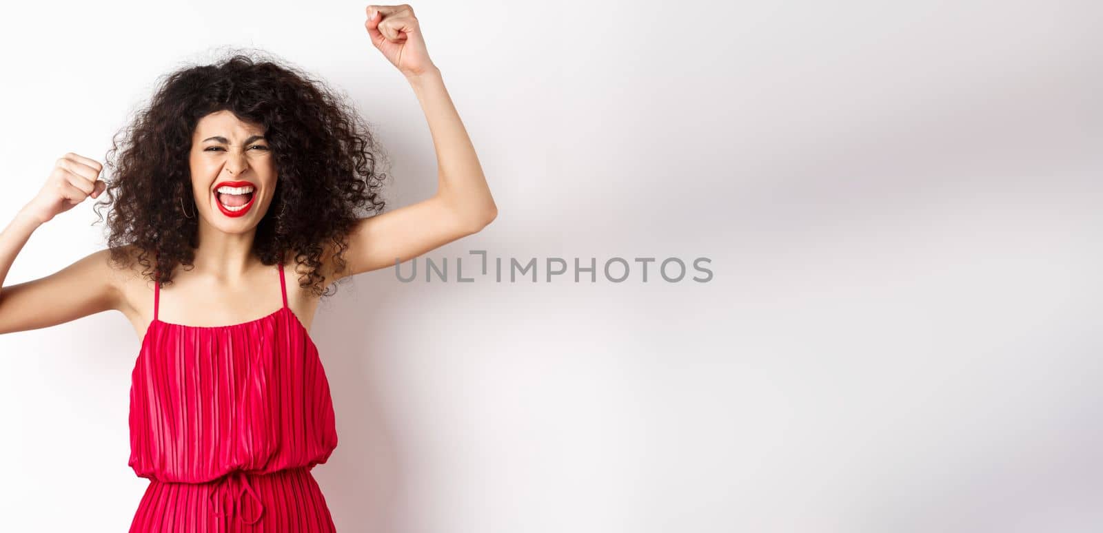 Cheerful emotive woman with curly hair, red dress, raising hands up and chanting, rooting for team, shouting wanting to win, standing on white background.
