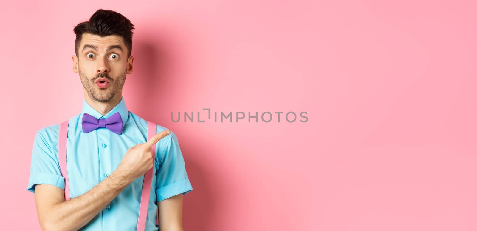 Intrigued and surprised cute guy with moustache showing awesome promo, looking curious and saying wow, pointing finger right at logo, standing on pink background.