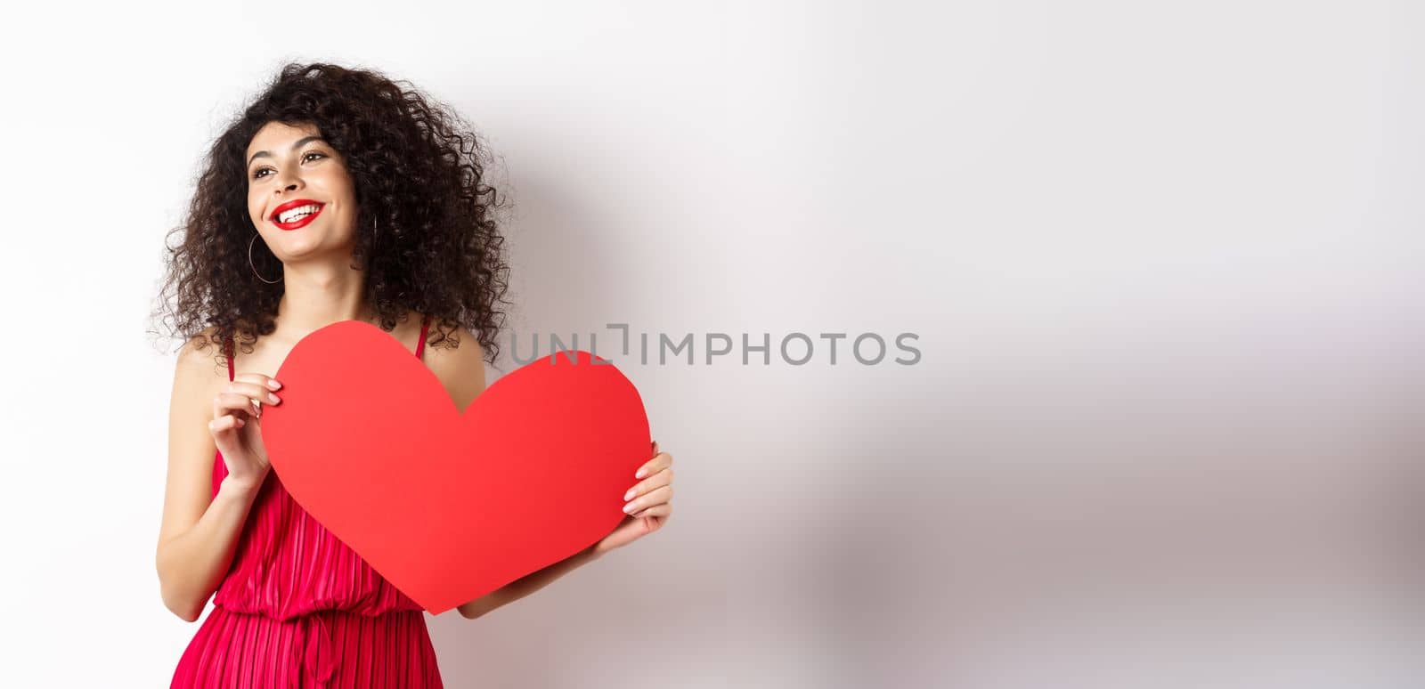 Romantic woman in dress and makeup, looking aside with dreamy face and love, showing big red valentines heart card, standing on white background by Benzoix