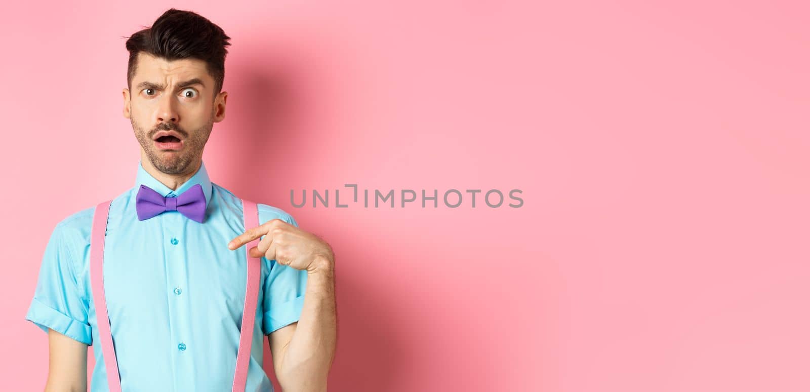 Image of confused funny guy in bow-tie and suspenders, pointing at himself as if being accused or chosen, raising eyebrow surprised, standing over pink background by Benzoix