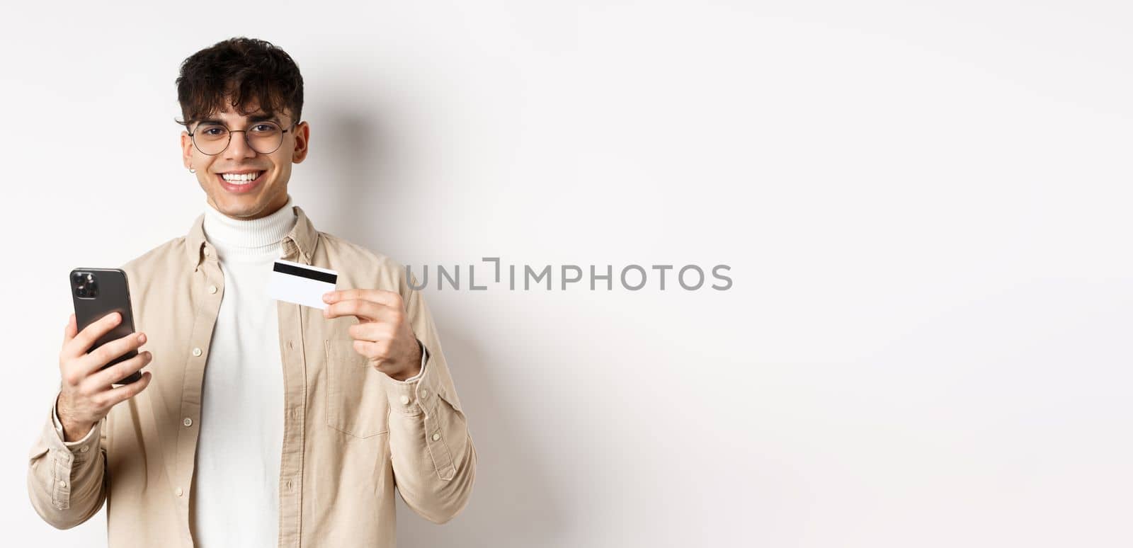 Portrait of natural young man in glasses paying in internet, showing smartphone and plastic credit card, standing on white background by Benzoix