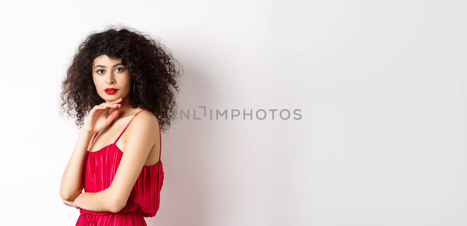 Beauty and fashion. Elegant lady with curly hair and red lips, fashionable makeup, wearing dress, gently touching chin and looking sensual at camera, white background by Benzoix