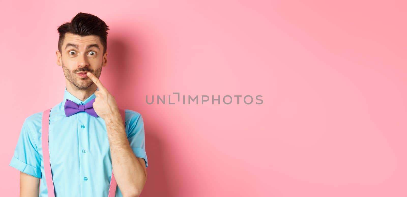 Intrigued happy guy touching lip, looking at camera pensive, making choice what to buy, standing over pink background.